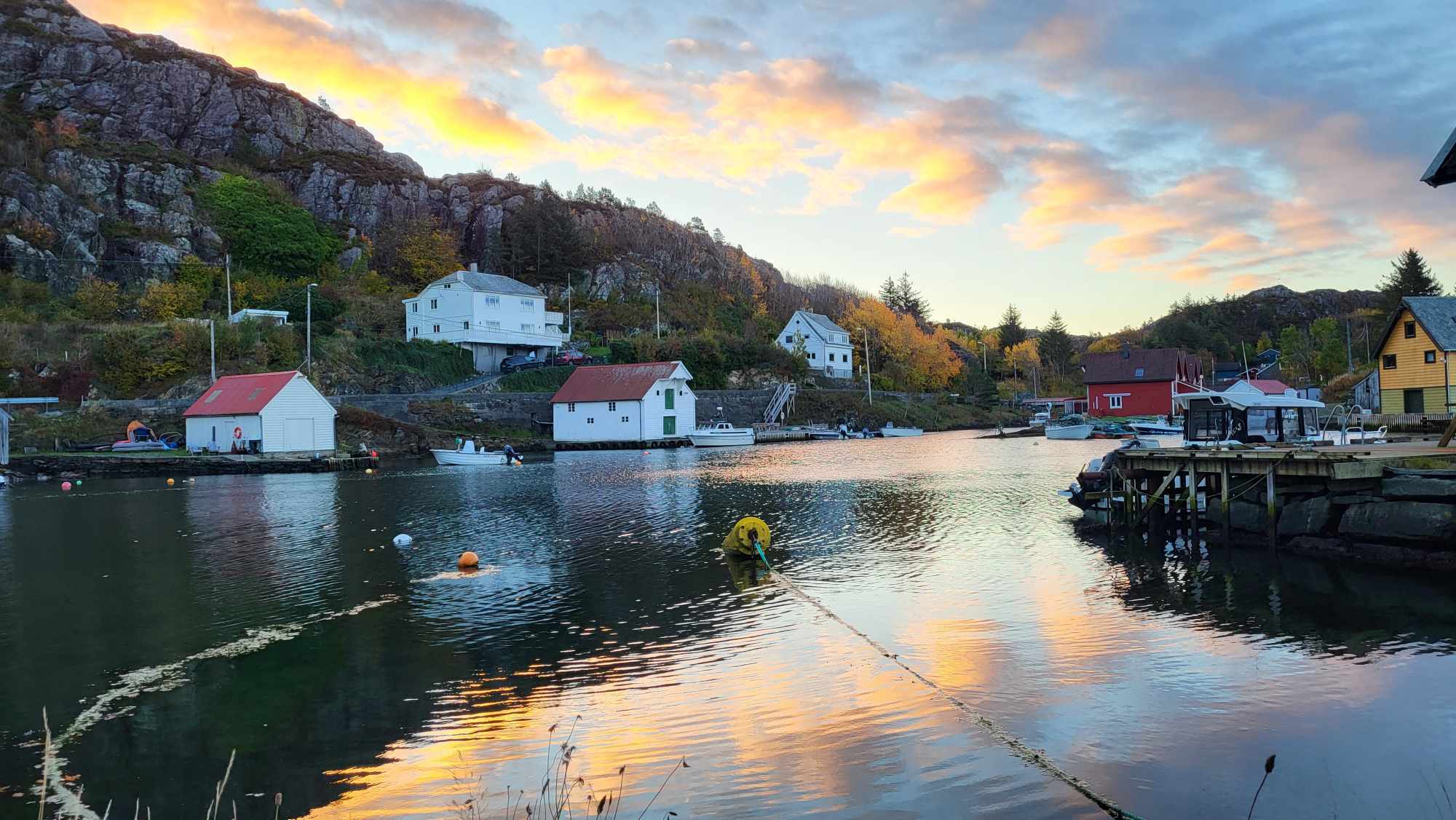 Lite kystmiljø med naust og hus ved vann, med speiling av solnedgang i sjøen.