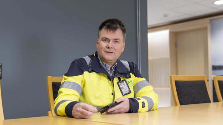 Mann i arbeidsjakke med refleks sitter ved bord og holder en mobiltelefon.